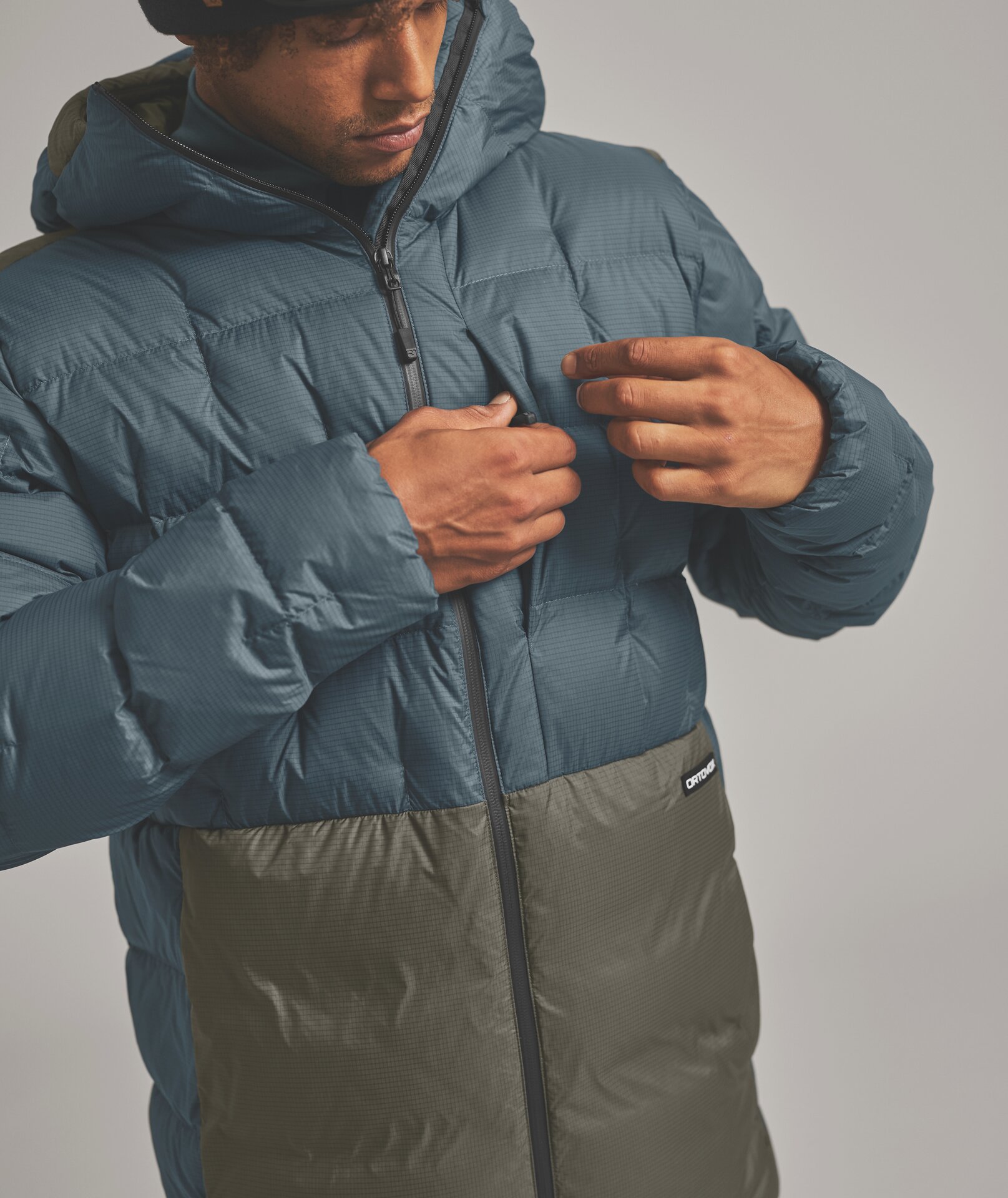 Person schliesst die Kapuzen-Daunenjacke; obere Hälfte blau, untere olive; Markenzeichen "ORTVOX" auf Hüfthöhe.