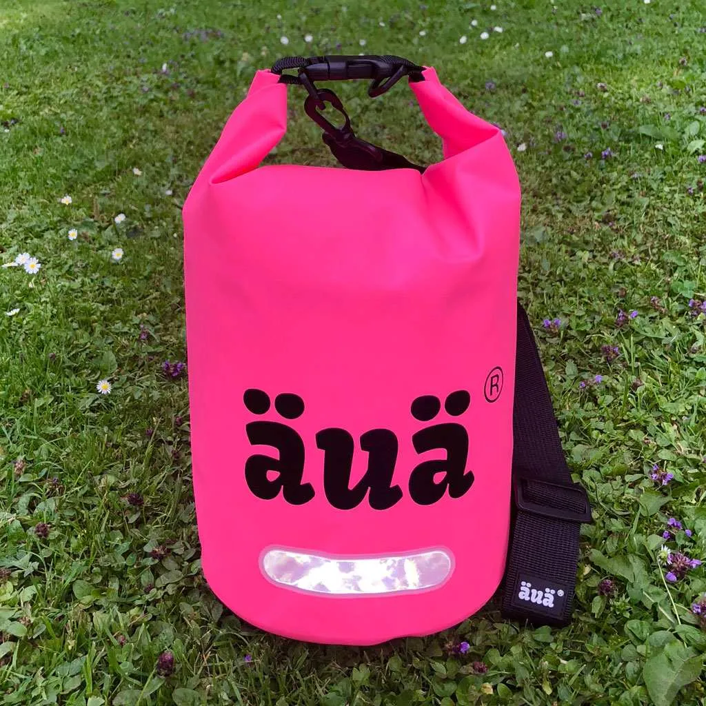 Pinker Dry Bag 5L auf grünem Rasen. Schwarze Tragegurte und reflektierendes Detail. "äua"-Logo in Schwarz.