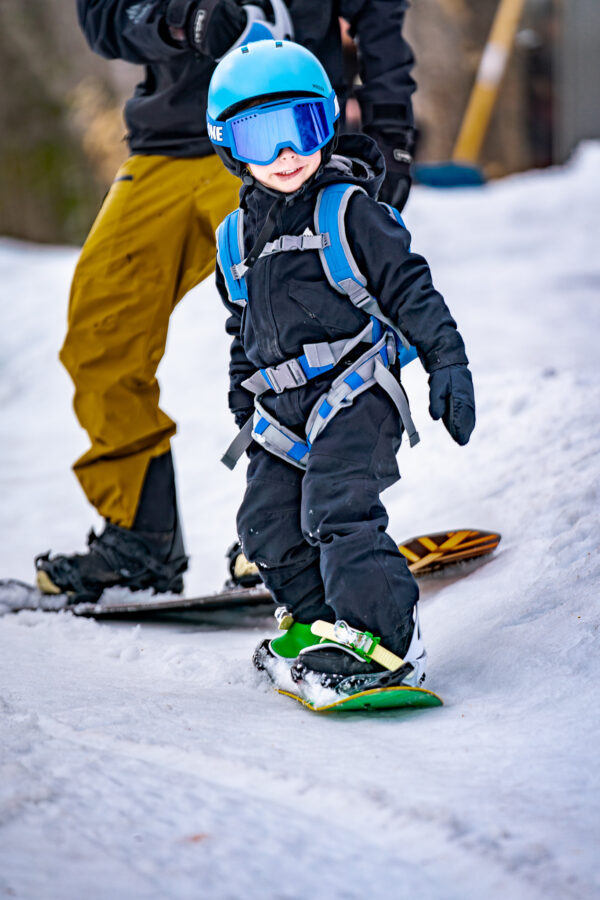 Un enfant portant un casque bleu et des lunettes de ski, équipé d'un harnais de snowboard « The One », apprend à faire du snowboard.
