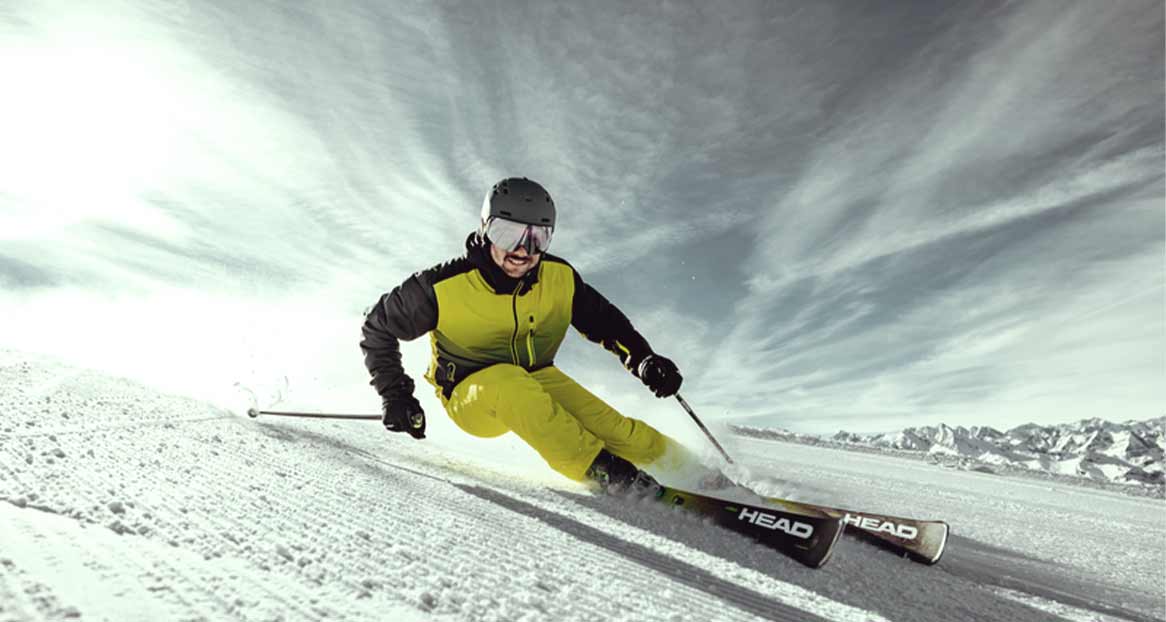 Skifahrer in gelber Kleidung und Helm fährt auf schneebedeckter Piste mit Bergen im Hintergrund.
