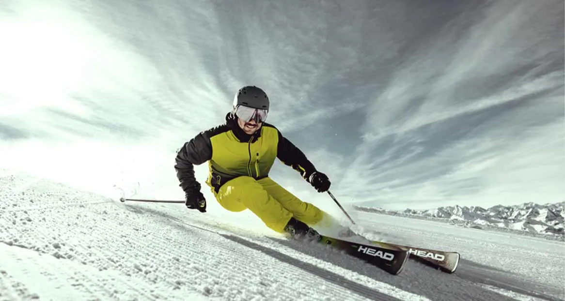 Skifahrer in gelber Kleidung und Helm fährt auf schneebedeckter Piste mit Bergen im Hintergrund.