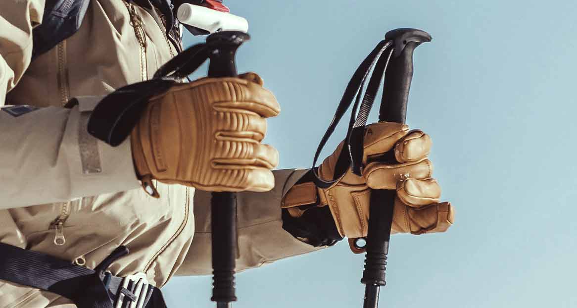 Deux mains gantées de cuir tiennent des bâtons de ski devant un ciel bleu clair. Une veste de ski beige est visible.
