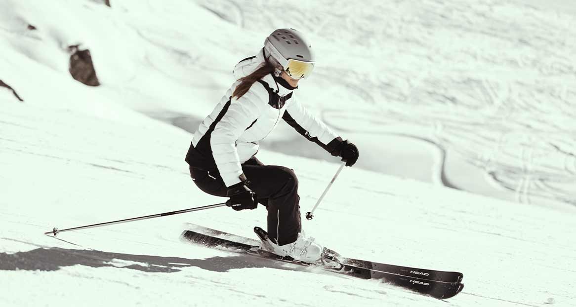 Eine Frau in Skikleidung fährt auf einer verschneiten Piste Ski. Sie trägt einen Helm und eine Brille.