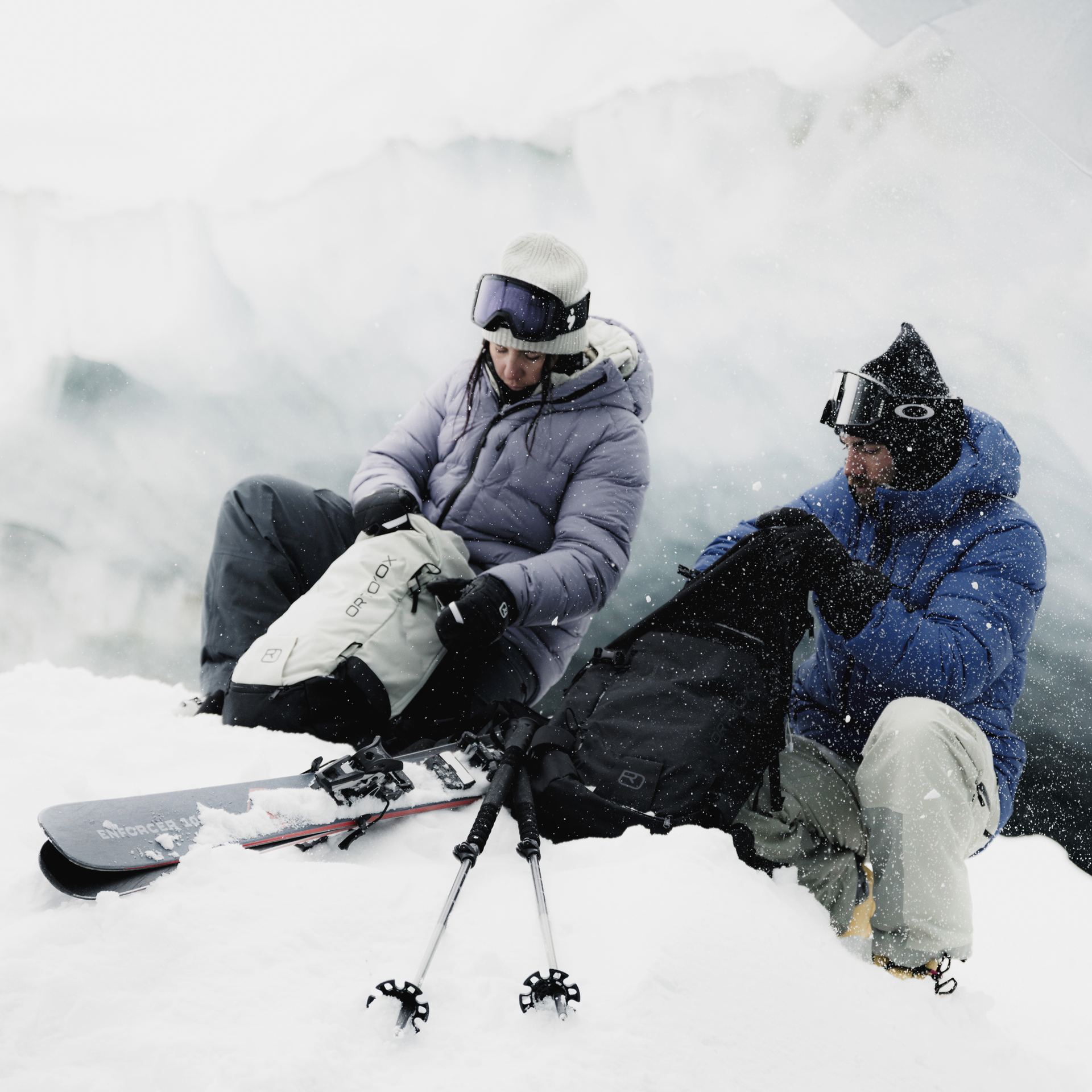 Zwei Skifahrer sitzen im Schnee vor einem Gletscher und öffnen ihre Rucksäcke. Skier und Stöcke liegen im Schnee.