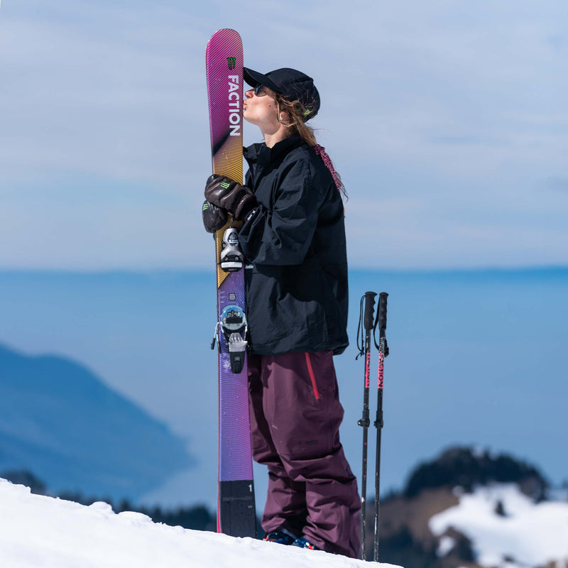 Frau mit Mütze küsst Ski auf einem schneebedeckten Gipfel, Skistöcke stehen daneben. Berge im Hintergrund.
