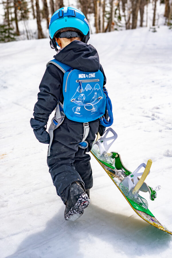 Petit enfant portant un casque bleu, un sac à dos et un snowboard sur une piste enneigée, vu de dos.