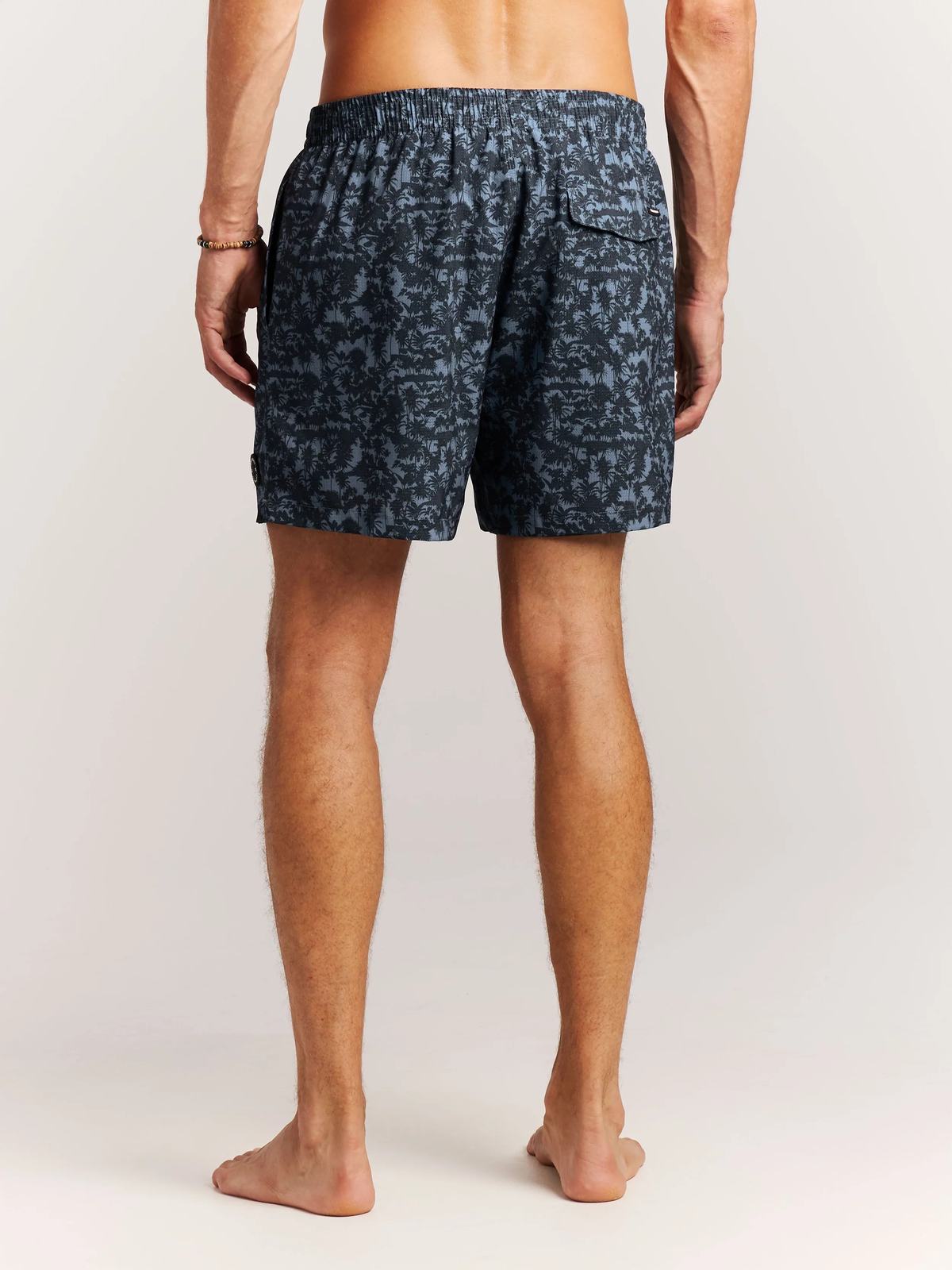 Ein Mann mit Badehose, von hinten gesehen, steht barfuß auf einem weißen Hintergrund. Seine Badehose ist blau mit dunklem Palmenmuster.