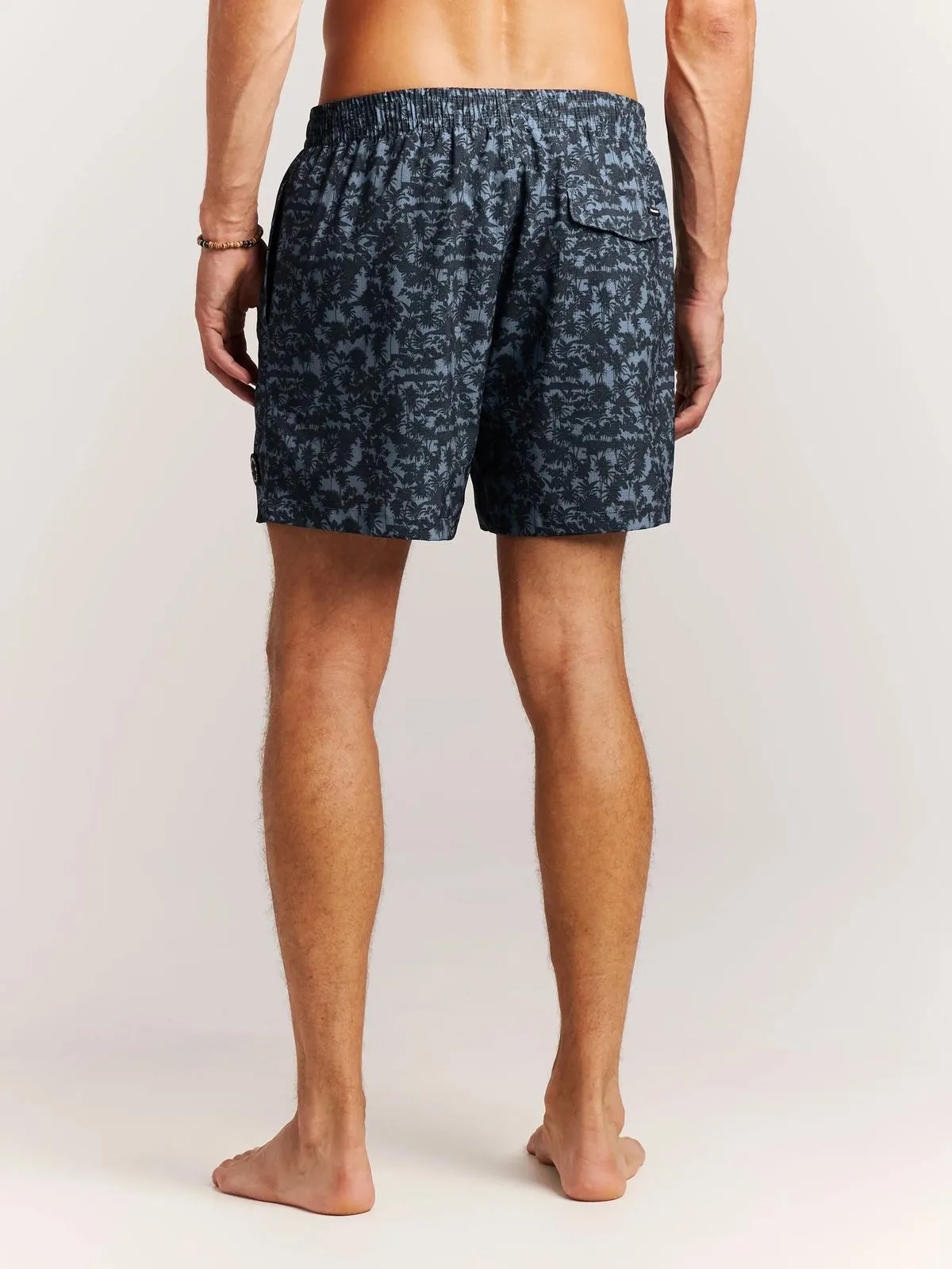 A man in swimming trunks, seen from behind, stands barefoot on a white background. His swimming trunks are blue with a dark palm tree pattern.