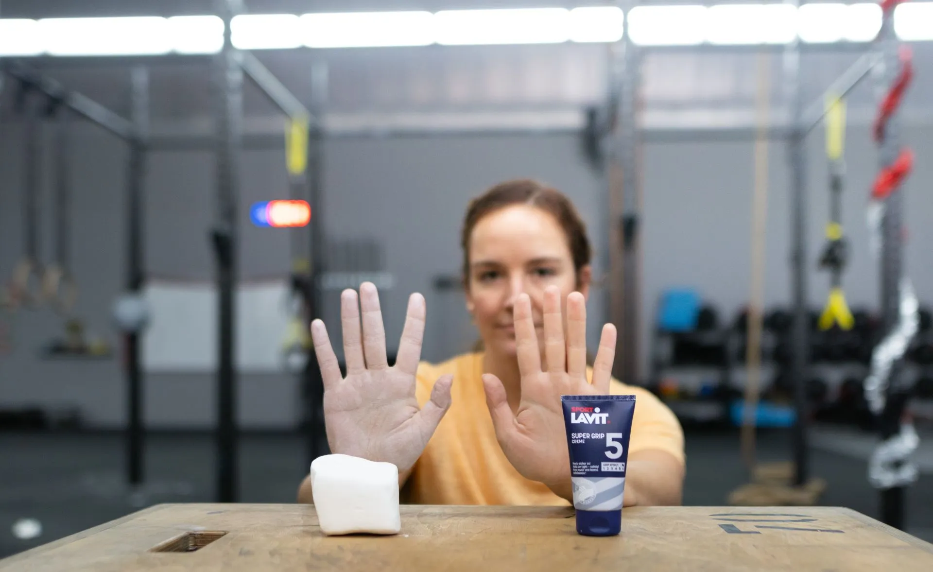 Frau zeigt Hände, Tube SPORT LAVIT Super Grip Creme und ein Magnesium Block auf Holztisch im Fitnessstudio.