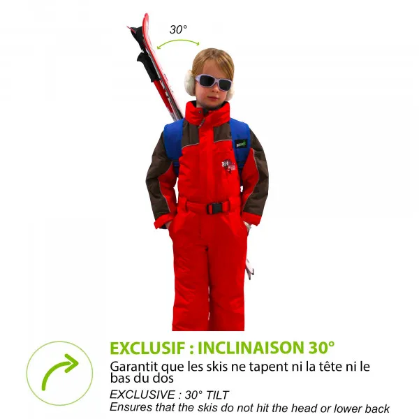 Enfant avec des skis sur le dos avec Skiback Kids Blue. Combinaison de ski rouge et lunettes de soleil. Les skis dépassent en biais.