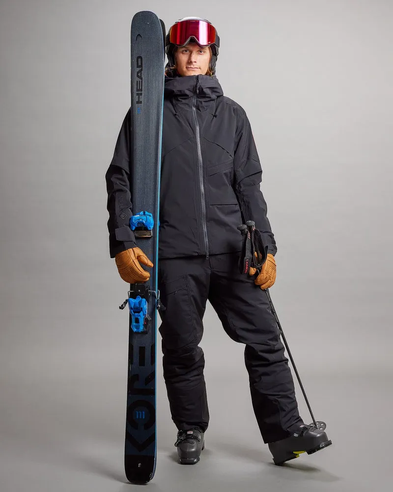 Person in schwarzer Gore-Tex Skihose und Jacke hält Ski und Stöcke, mit roter Skibrille und Handschuhen.