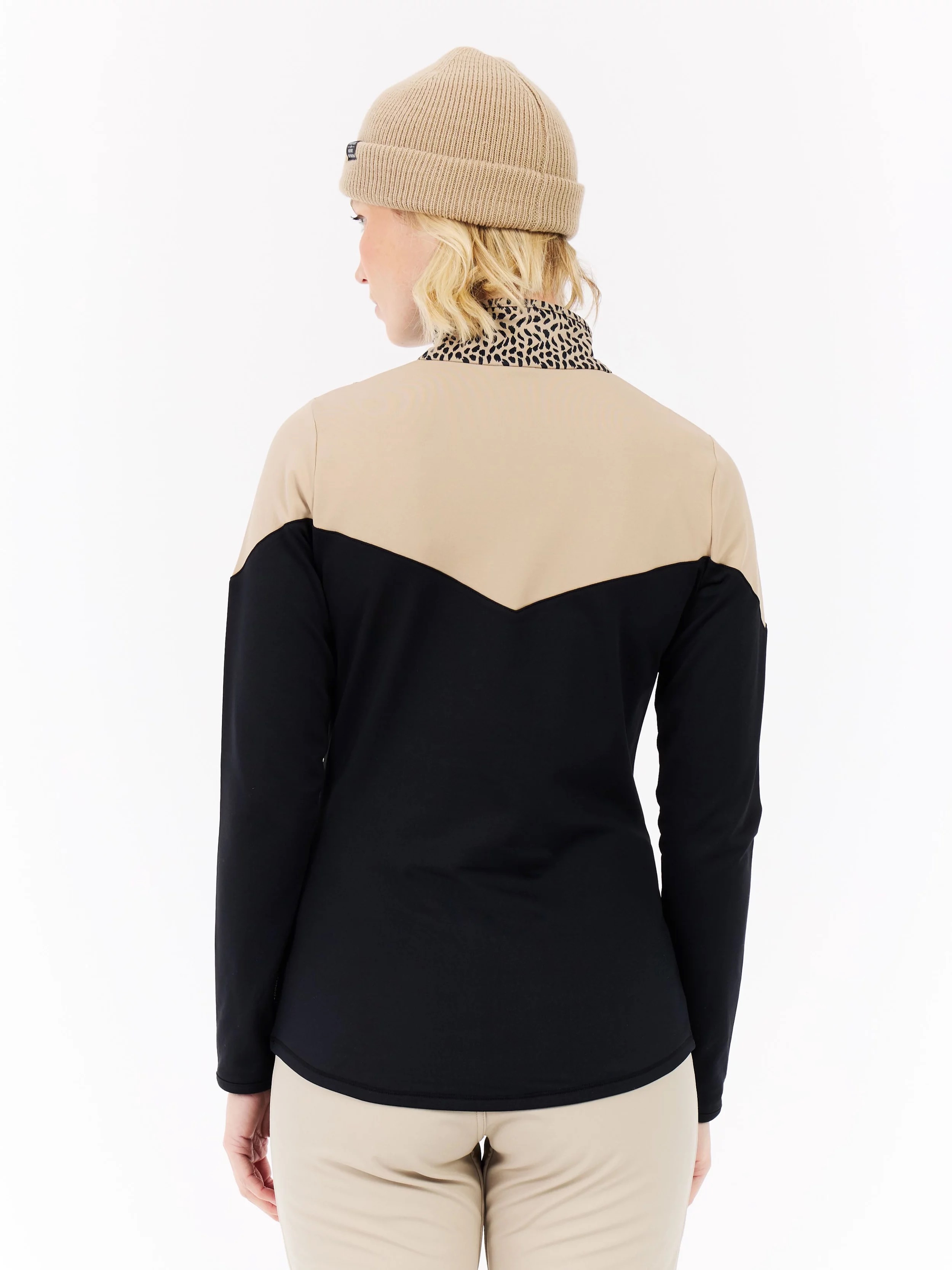 Rear view of a woman wearing a beige cap, a beige, v-shaped jacket over a black top and trousers in a matching color.