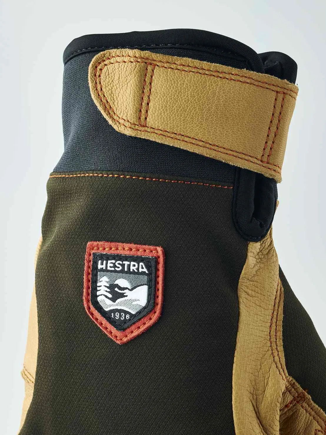 Détail d'un gant avec dessus marron, détails en cuir et logo "Hestra 1936". Le gant a une manchette avec fermeture velcro.