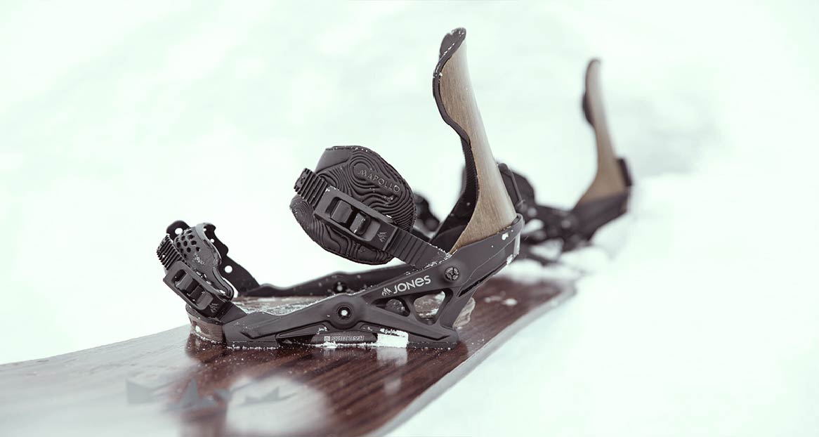 Gros plan d'une fixation de snowboard de Jones sur un snowboard dans la neige. La fixation est noire avec des accents bruns.