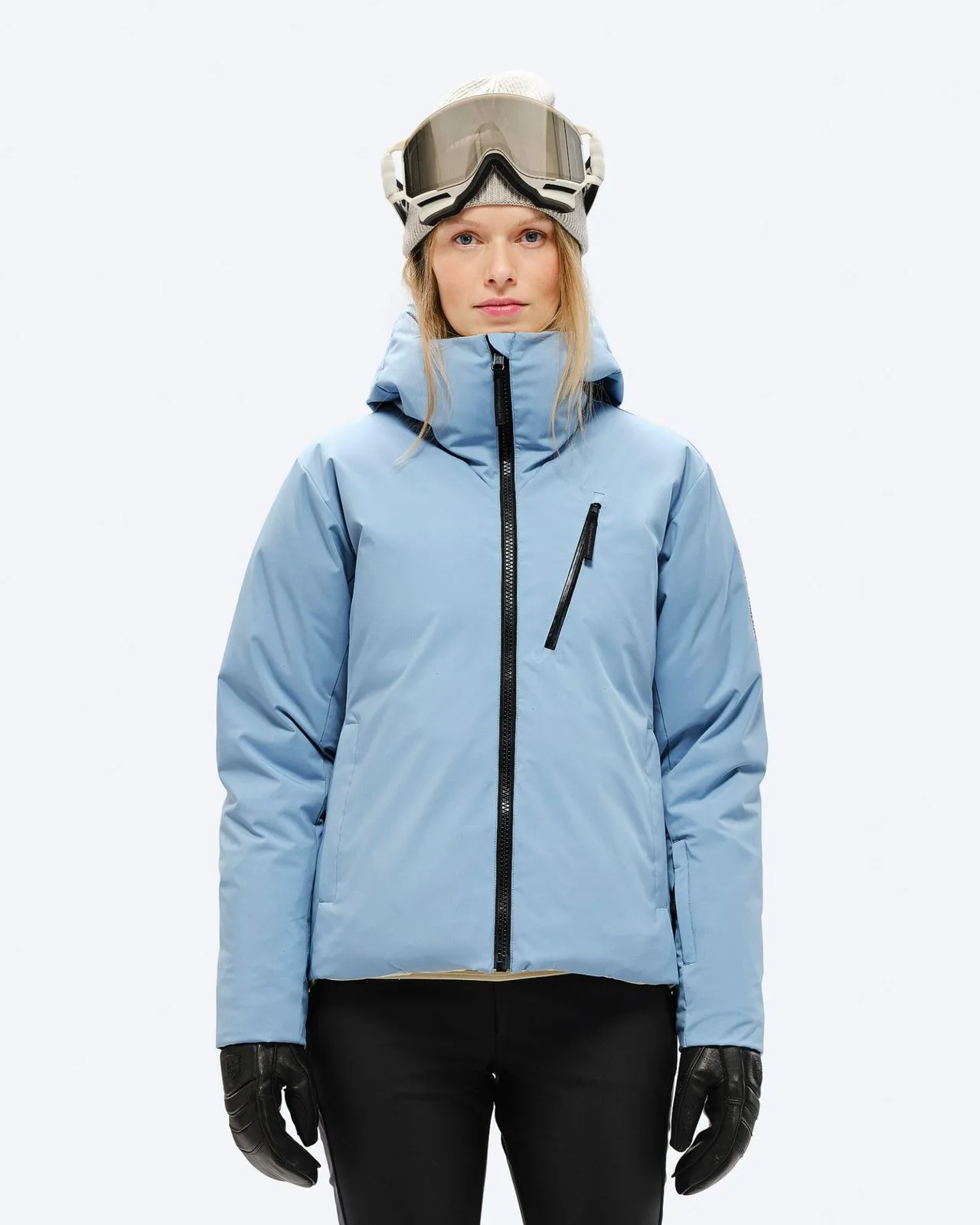 A woman with a cap and ski goggles wears a light blue down jacket with a hood and black zipper.