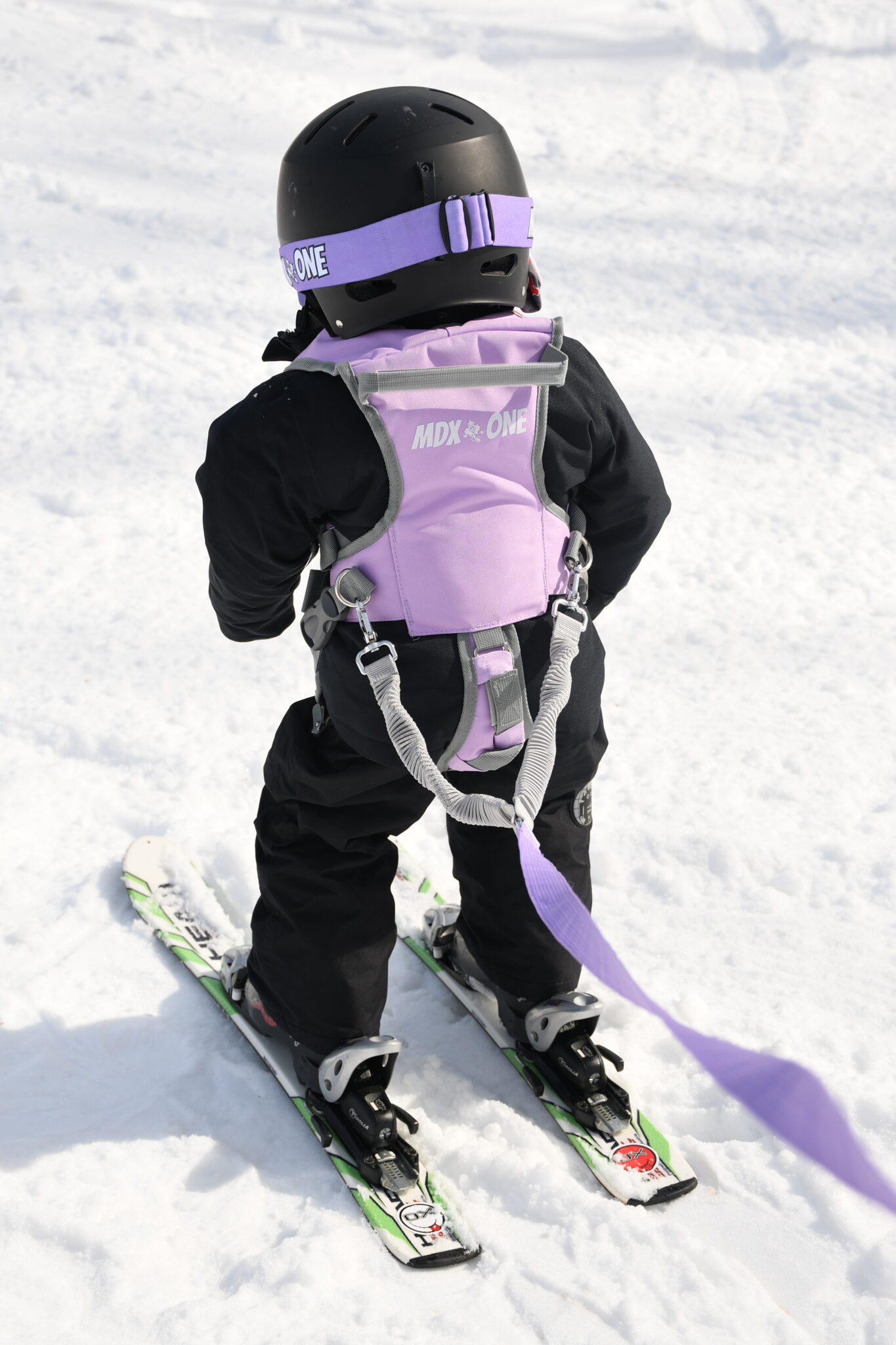 Child with ski equipment, black helmet, purple harness and leash, standing on snow.