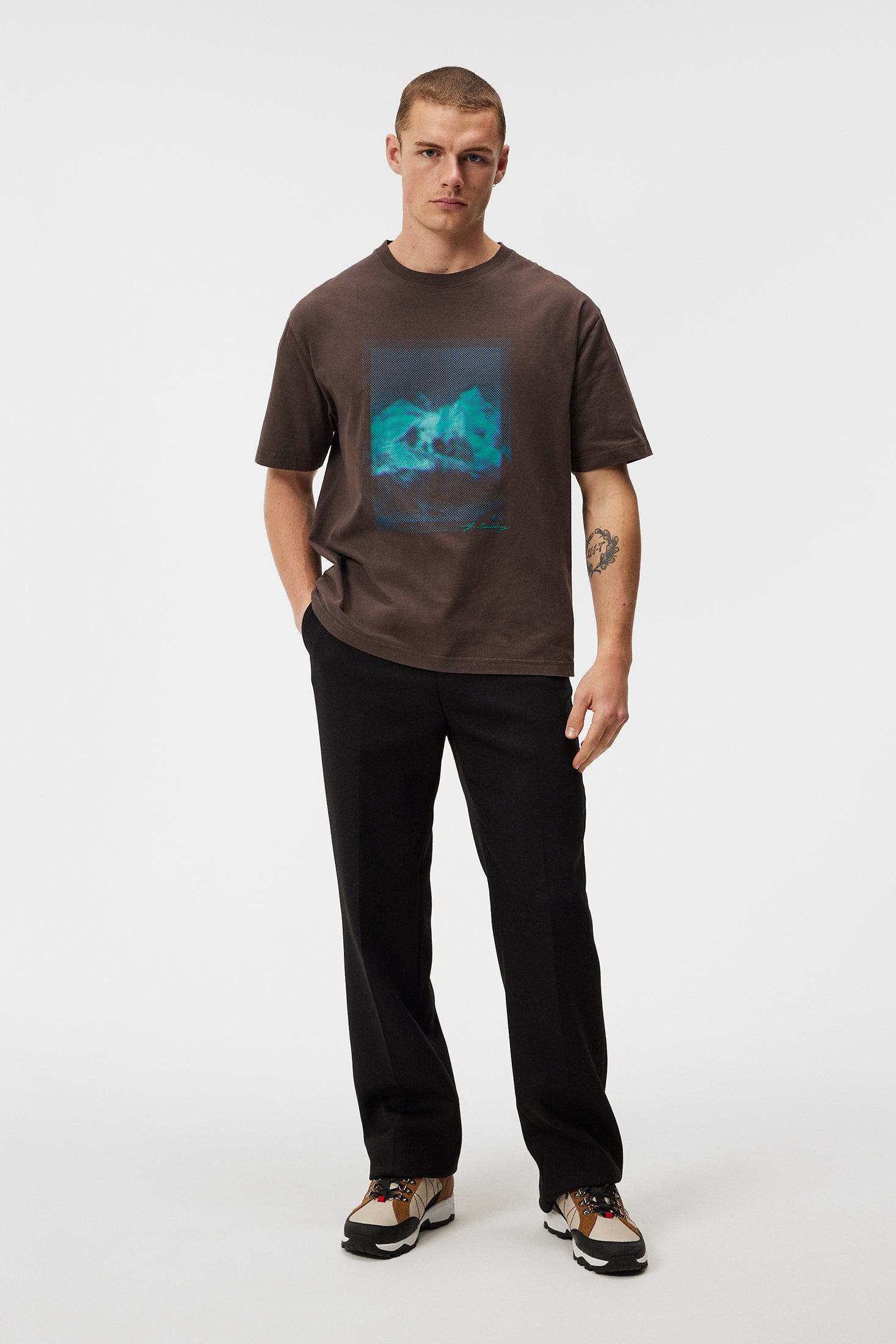 Mann trägt braunes T-Shirt mit blauem Print, schwarze Hose und Sneakers vor weißem Hintergrund.
