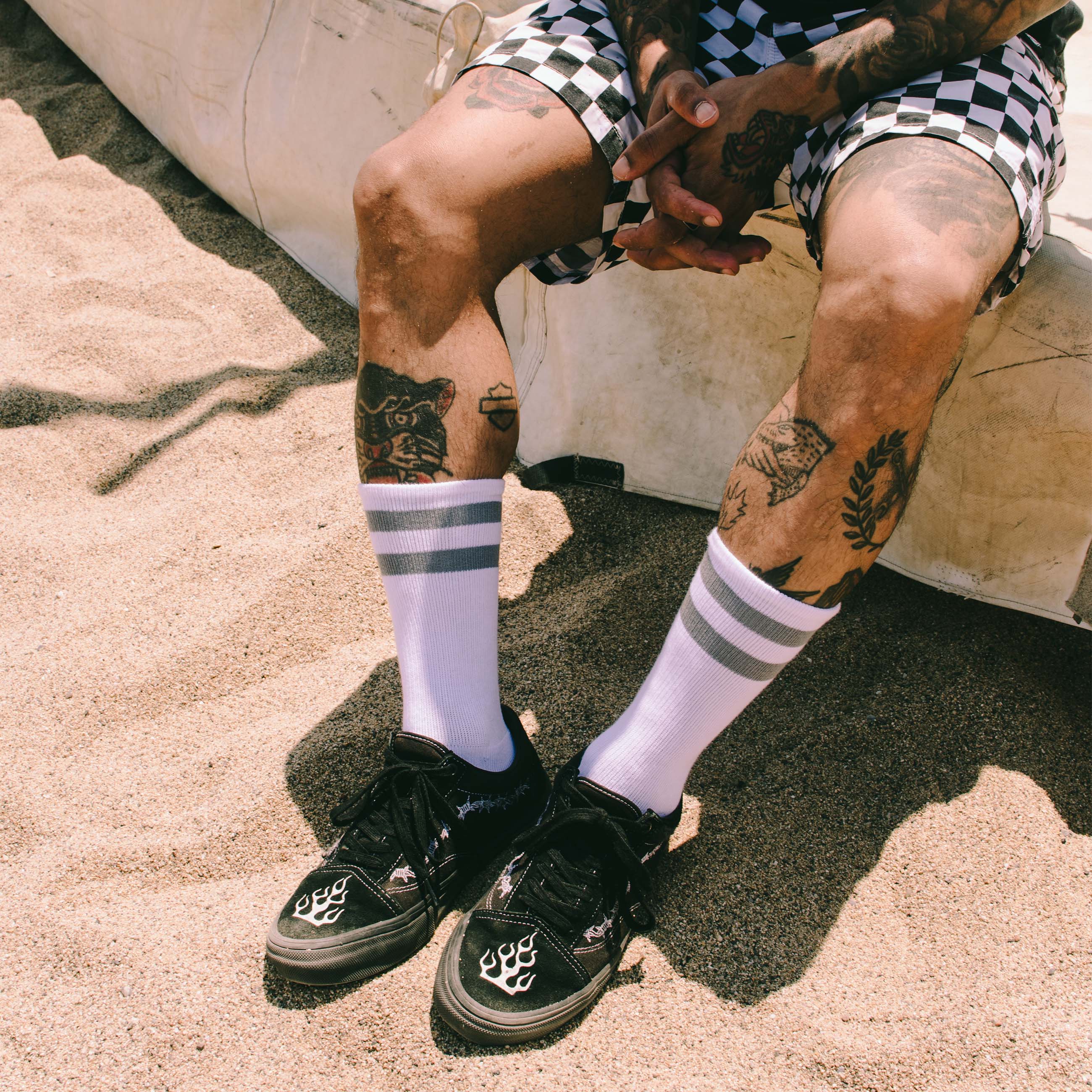 Strandlook mit Tattoos Person mit Tattoos trägt Schachbrettmuster-Shorts, Falkor Mid High Socken, und Turnschuhe am Strand.