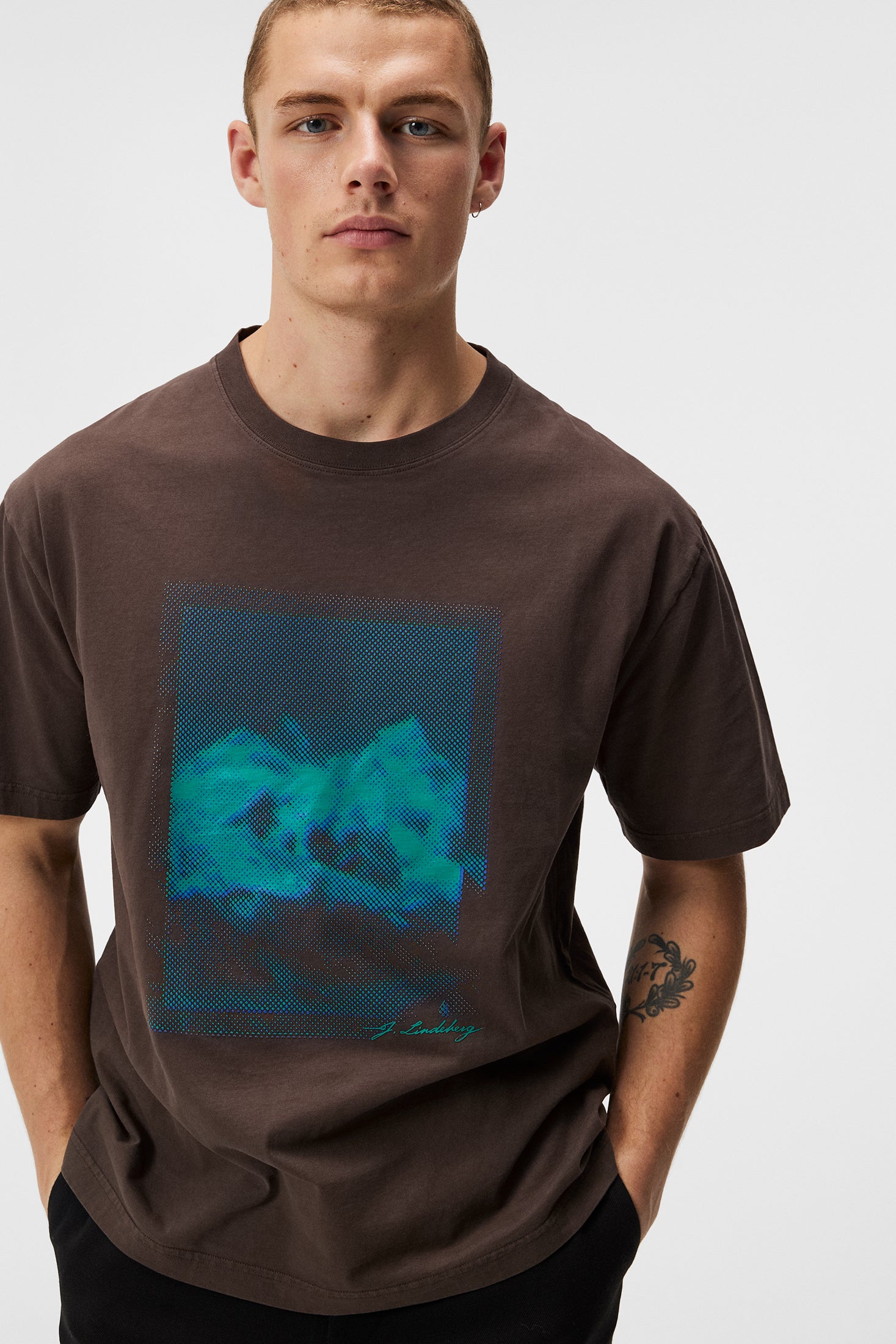 Mann trägt braunes T-Shirt mit blau-grünem Print. Der Print ist ein abstraktes Muster mit dem Schriftzug "J. Lindeberg".