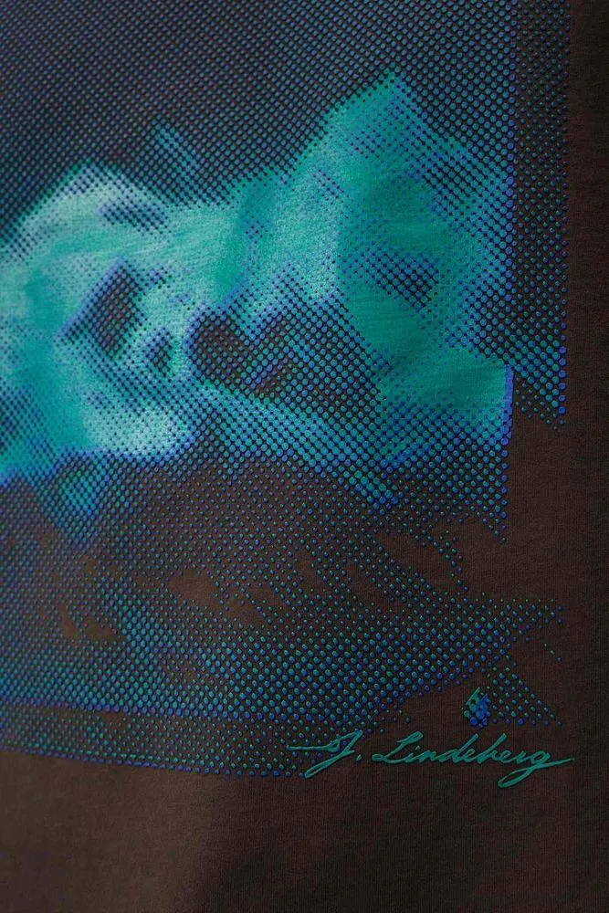 Close-up of the Walsh Boxy Print Tee Delicioso with blue-green seasonal print and J.Lindeberg lettering on brown fabric.