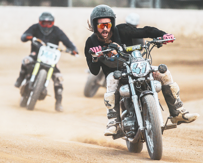 Motorradrennfahrer 47 Motorradrennen: Fahrer mit rosa-grauer Sonnenbrille, Nummer 47, Staub wirbelt auf.
