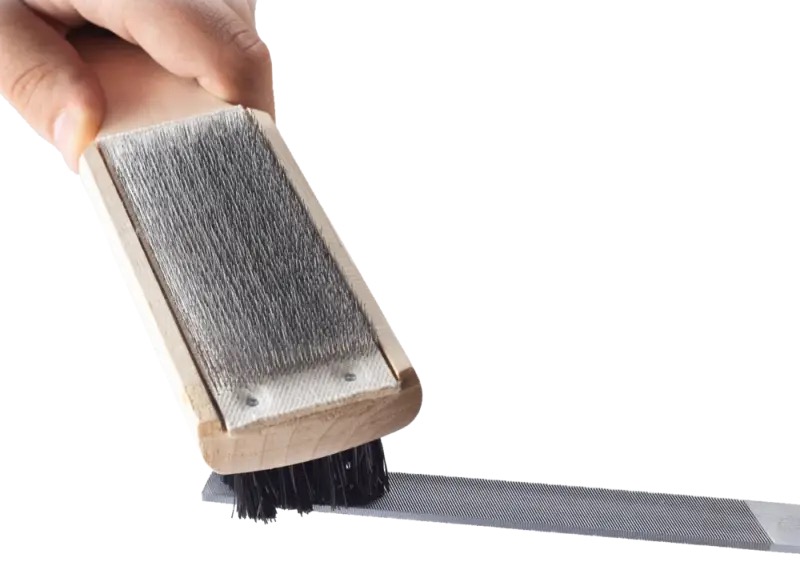 Spazzola per lime bifacciale Una mano pulisce una lima grossolana con una spazzola per lime double-face. La spazzola ha setole metalliche su un lato e setole di nylon sull'altro.