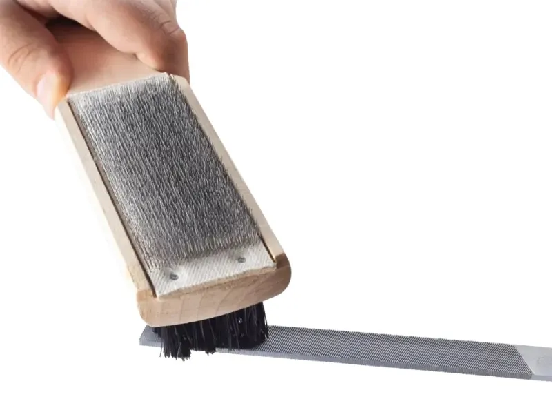 One hand cleans a coarse file with a double-sided file brush. The brush has metal bristles on one side and nylon bristles on the other.