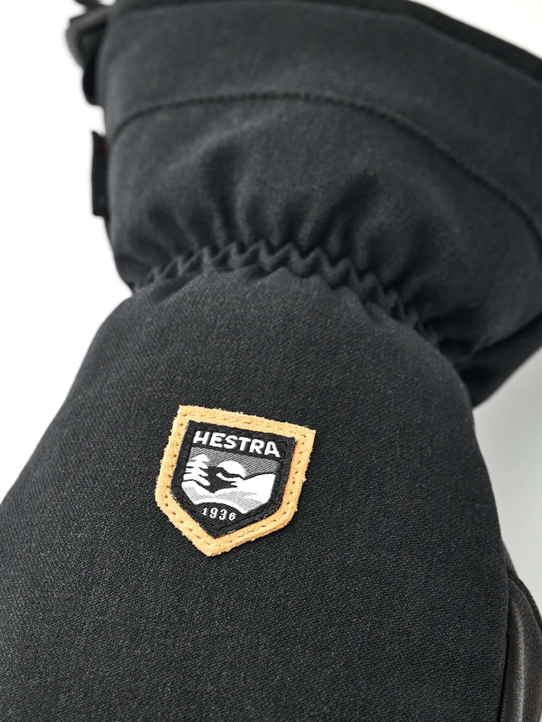Détail d'une moufle noire avec le logo Hestra, une plaque orange bordée d'un motif de montagne et l'année de fondation 1936.