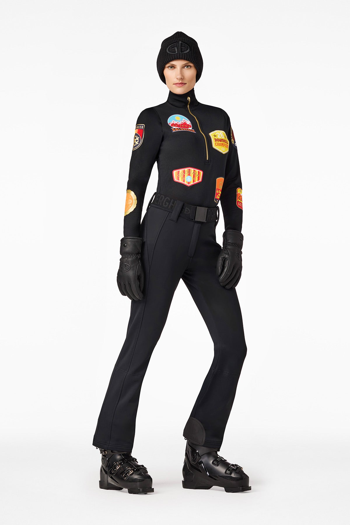 Woman in black ski clothing with patches, gloves, hat and ski boots against a white background.