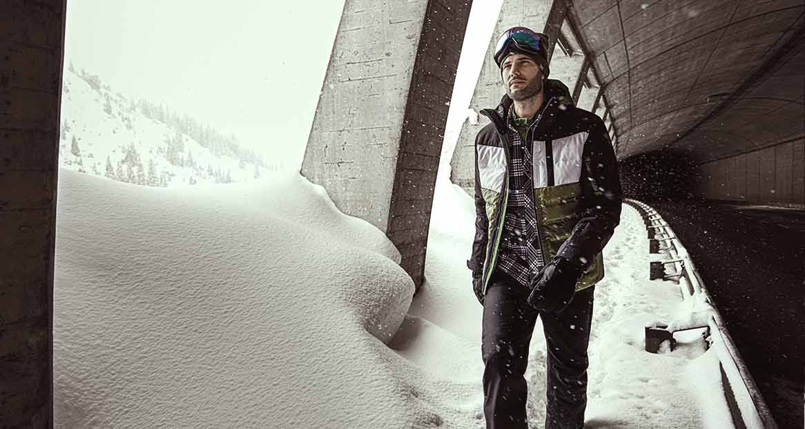 Ein Mann mit Skibrille, Winterjacke, Handschuhen und Hose steht an einem schneebedeckten Ort.