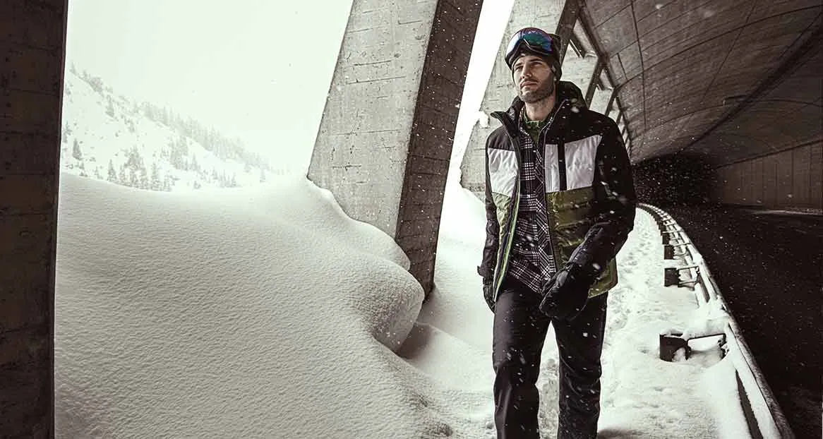 Ein Mann mit Skibrille, Winterjacke, Handschuhen und Hose steht an einem schneebedeckten Ort.