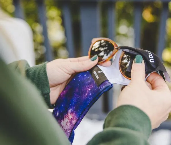 Hände legen Sonnenbrille in einen "Galactic Sunnysoc" Brillenputztuch, Hülle mit Galaxie-Muster.