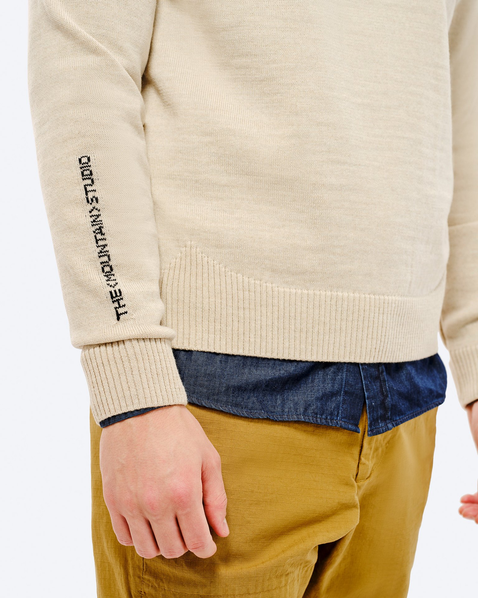 St. Moritz Ski Sweater Detailed view of the St. Moritz Ski Sweater, beige, with lettering on the sleeve, over a blue shirt and beige pants.