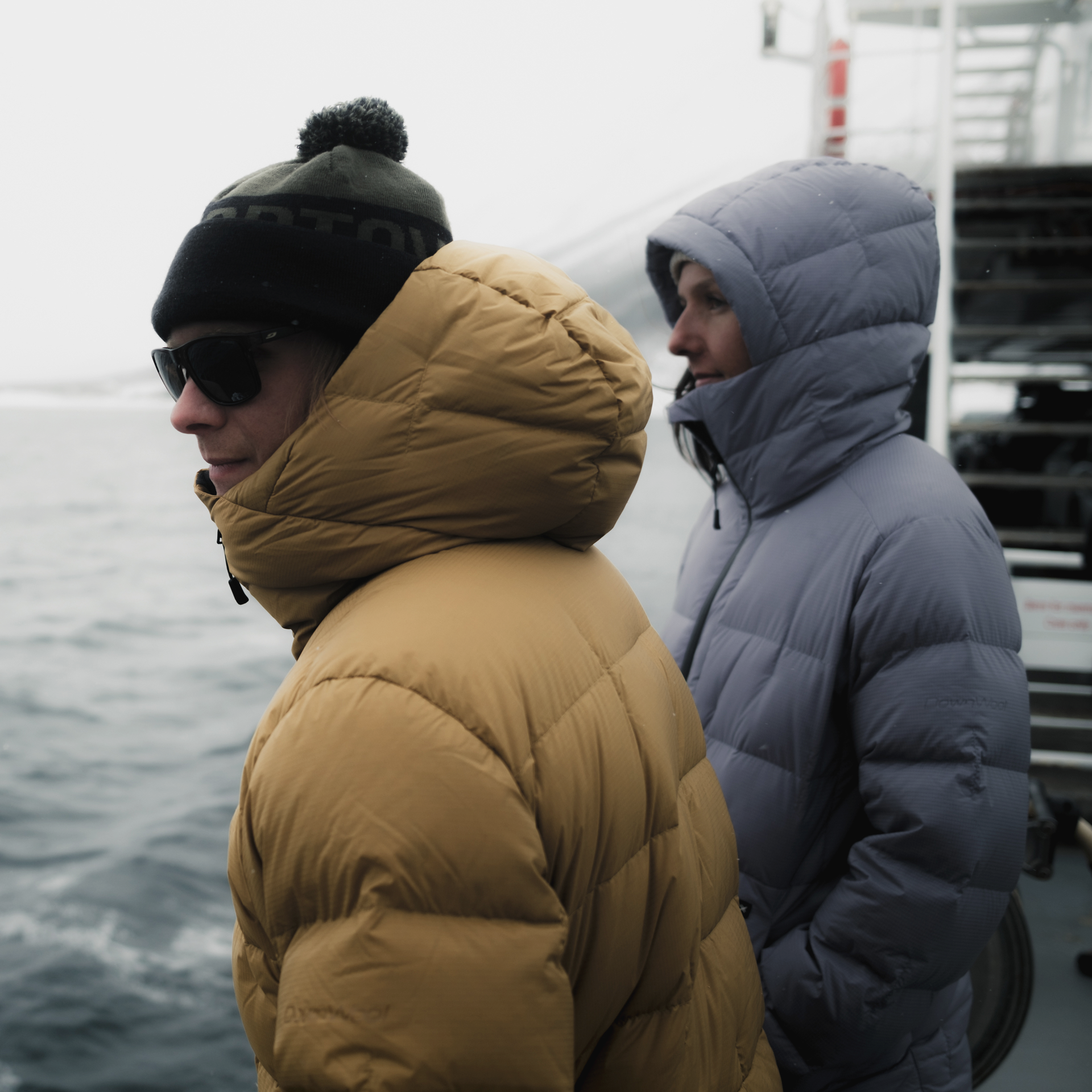 Zwei Personen, in Winterkleidung gehüllt, stehen an Deck eines Schiffes und blicken aufs Meer.