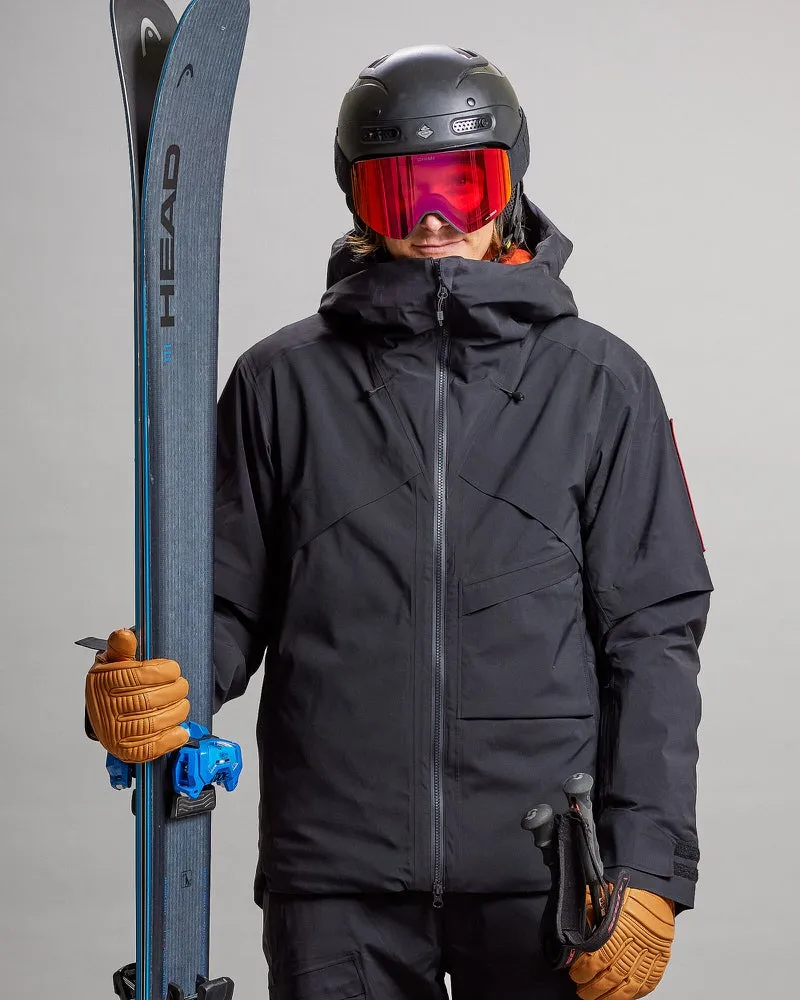 Person in schwarzer Gore-Tex-Skijacke mit Ski, Helm und Brille, hält Stöcke und trägt Handschuhe.