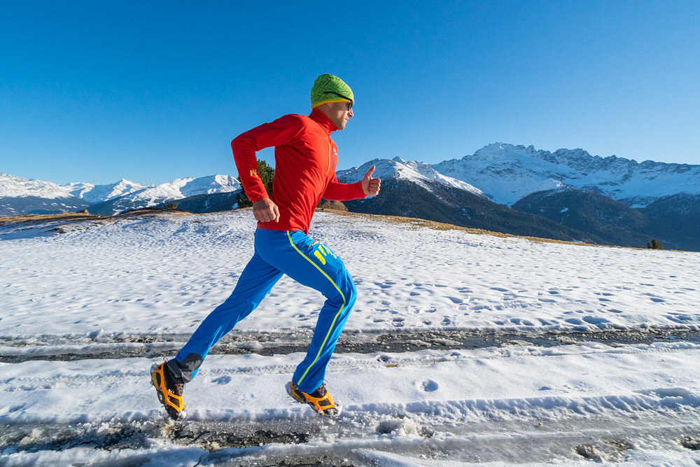 Un homme au pas de course avec des crampons de chaussures Trail 2.1 sur une surface enneigée devant un panorama de montagnes.