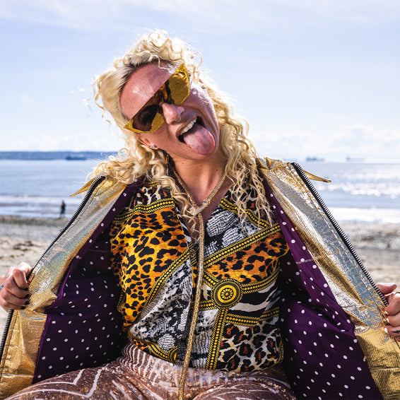 Personne blonde avec des lunettes de soleil, langue sortie, portant une veste avec des accents de couleur pyrite.