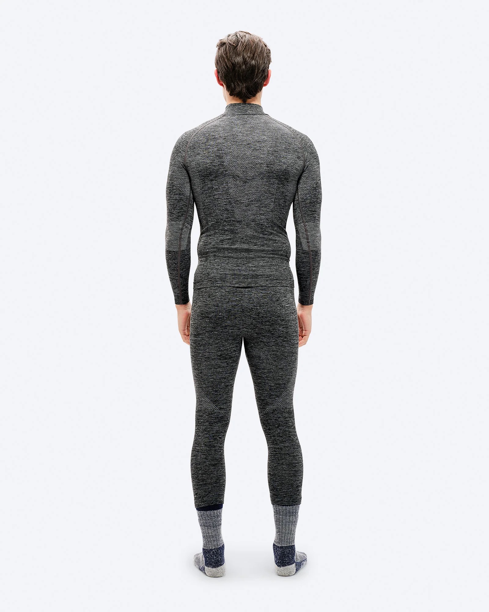 A man from behind, dressed in a gray, tight-fitting, long-sleeved thermal shirt and trousers, wearing thick socks. The background is white.