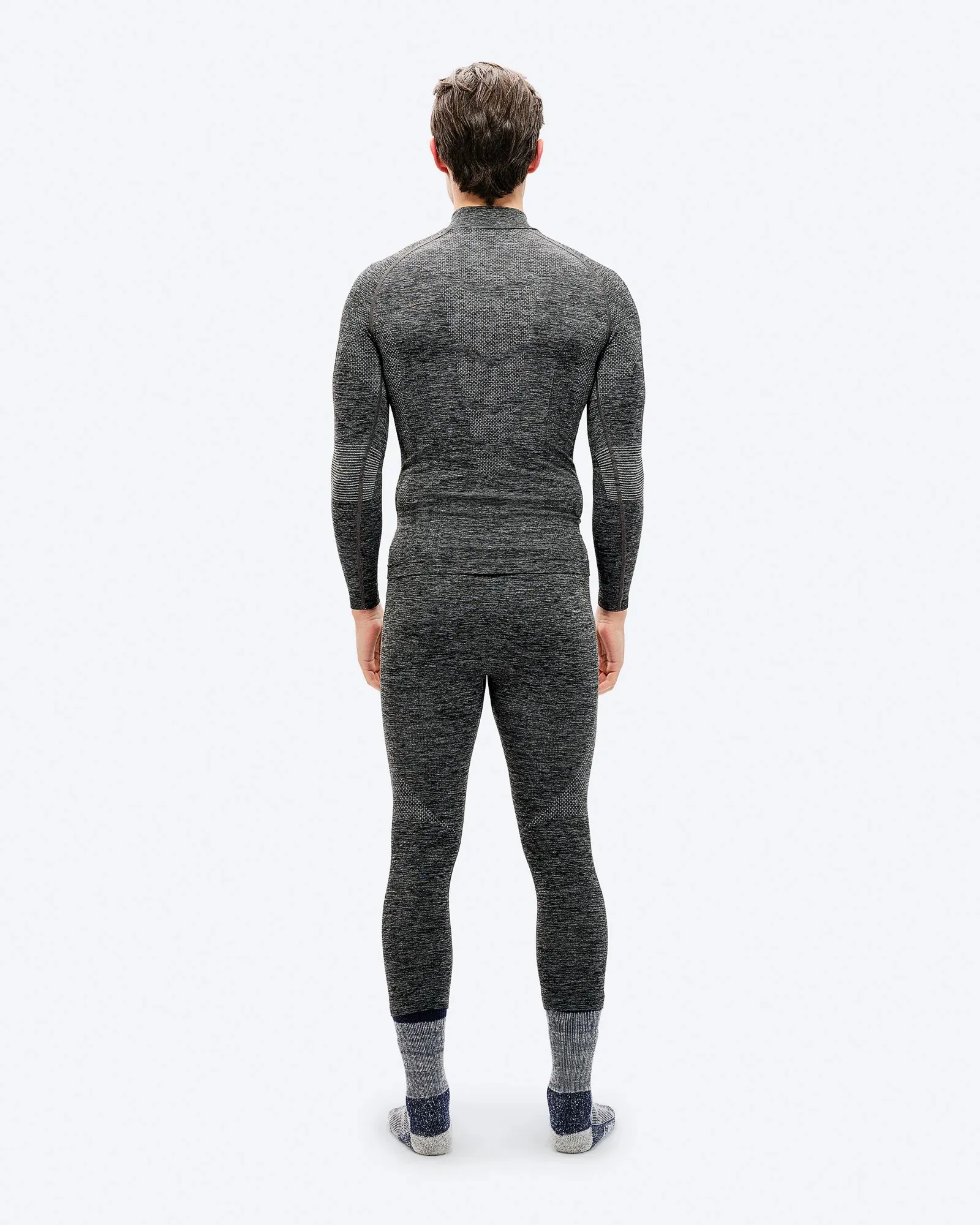 Ein Mann von hinten, gekleidet in ein graues, eng anliegendes, langärmeliges Thermoshirt und eine Hose, trägt dicke Socken. Der Hintergrund ist weiß.
