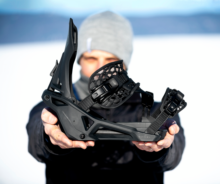 Schwarze Snowboardbindung Eine Person hält eine schwarze Snowboardbindung in die Kamera, im Hintergrund ist Schnee zu sehen.