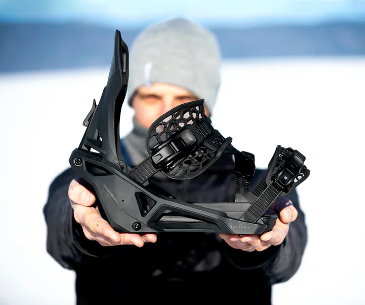 Una persona tiene un attacco da snowboard nero davanti alla telecamera, sullo sfondo si vede la neve.