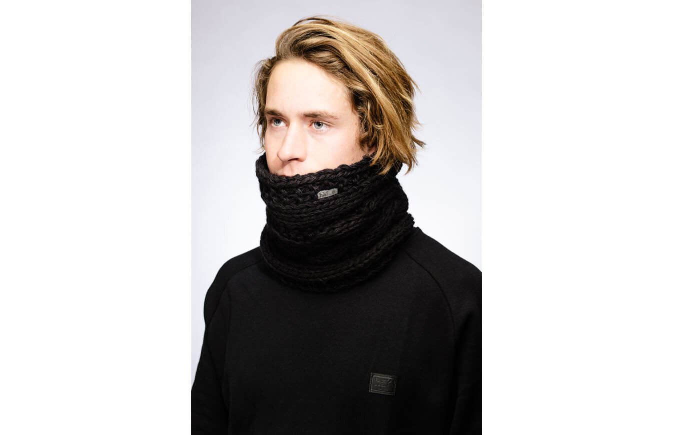 Man with blond hair wearing a black knitted stroller neckwarmer. Logo patch on the neck and sweater.