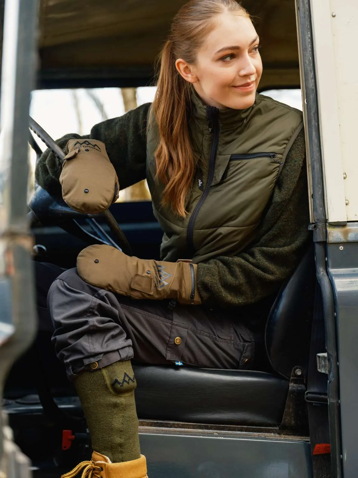 Donna in auto con calze da riscaldamento. Calzettoni verdi, scarpe marroni, gilet verde oliva, guanti beige.