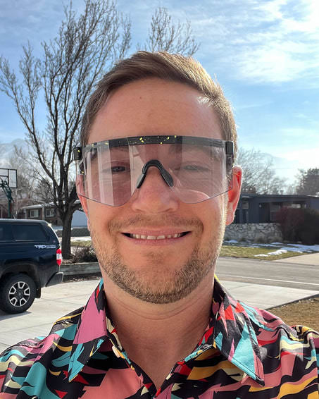 Ein Mann lächelt und trägt eine durchsichtige Cosmos Photochromic 2000 Sonnenbrille mit bunten Akzenten auf dem Rahmen.