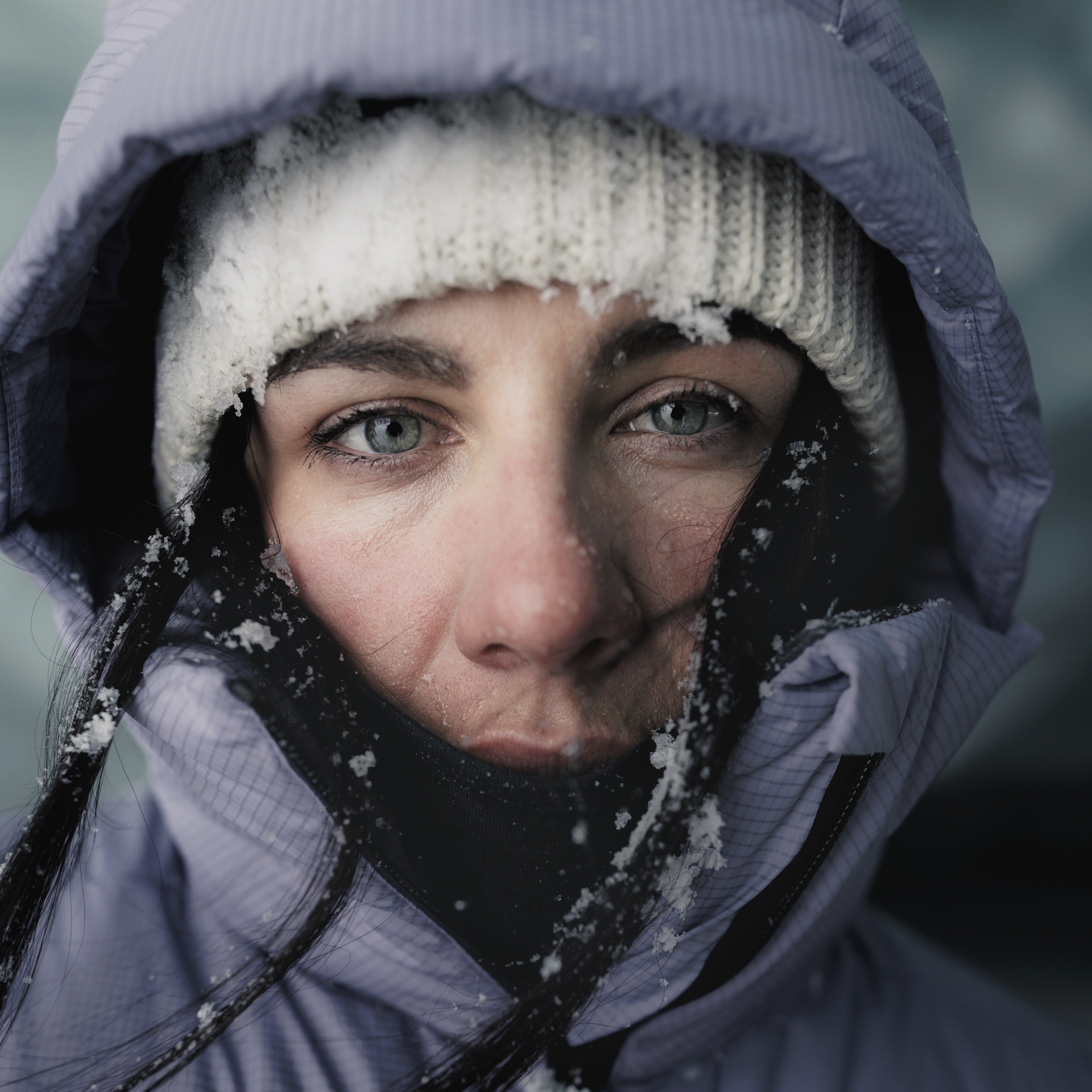 Nahaufnahme einer Frau mit eisbedeckter Mütze und Kapuze, die direkt in die Kamera blickt. Sie hat grüne Augen und dunkle Haare.