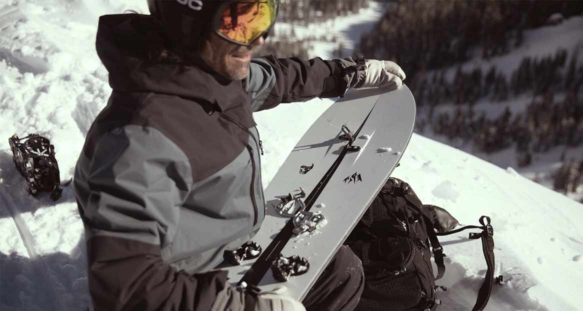 Homme avec un splitboard dans un paysage de montagne enneigé. Il porte un casque, des lunettes de ski et des vêtements d'hiver. Sac à dos et fixations dans la neige.