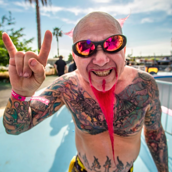 Mann mit Tattoos, Sonnenbrille, pinkem Bart und Hörnern zeigt Rocker-Geste. "The Combustion" Produkt.
