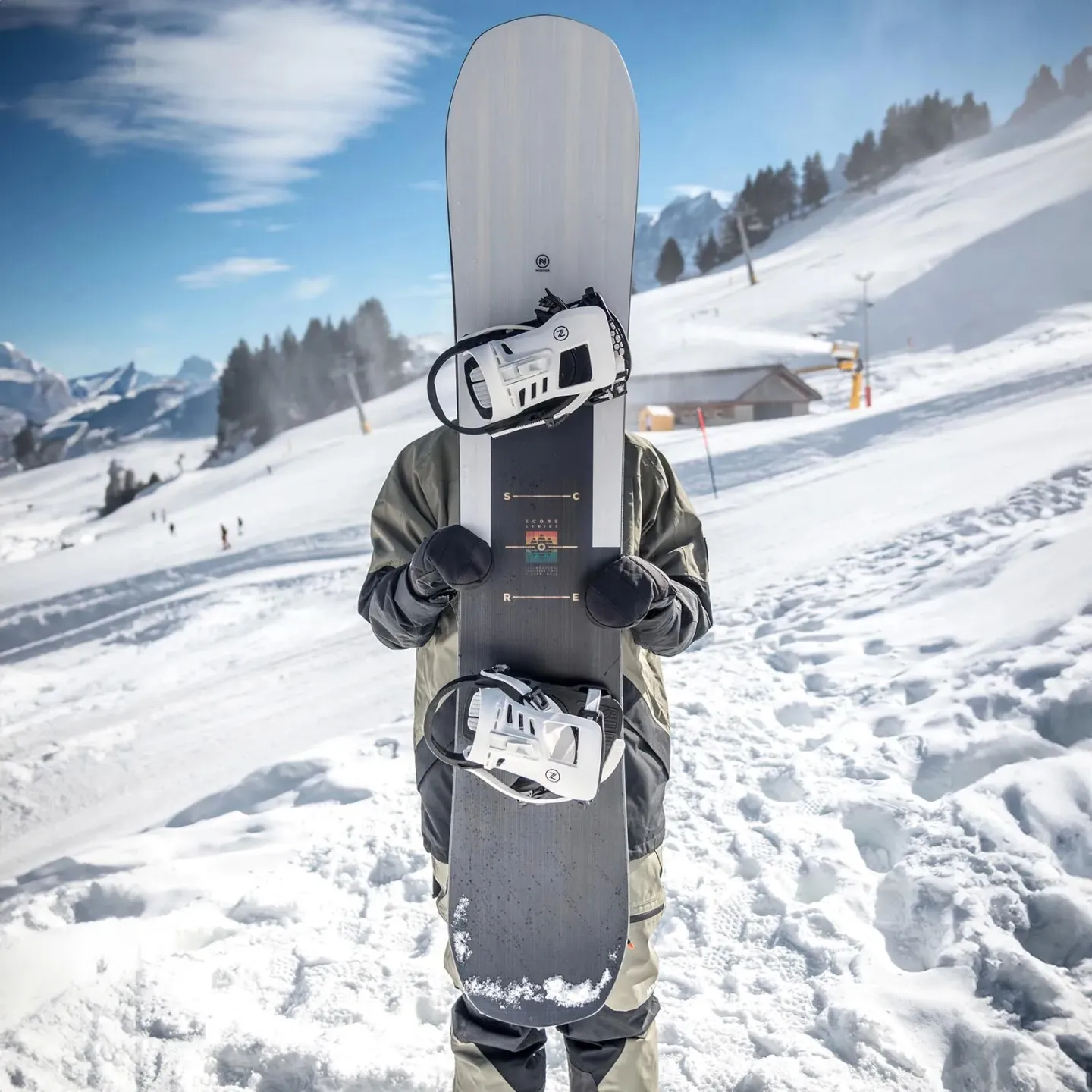 Person hält ein Nidecker Score Snowboard mit montierten Bindungen vor dem Körper; verschneite Bergkulisse.