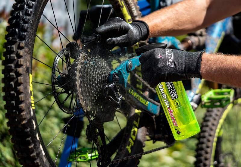 Reinigung einer Fahrradkette mit Muc-Off Drivetrain Cleaner und Bürste, Handschuhe, Kettenreiniger Sprühflasche im Fokus.
