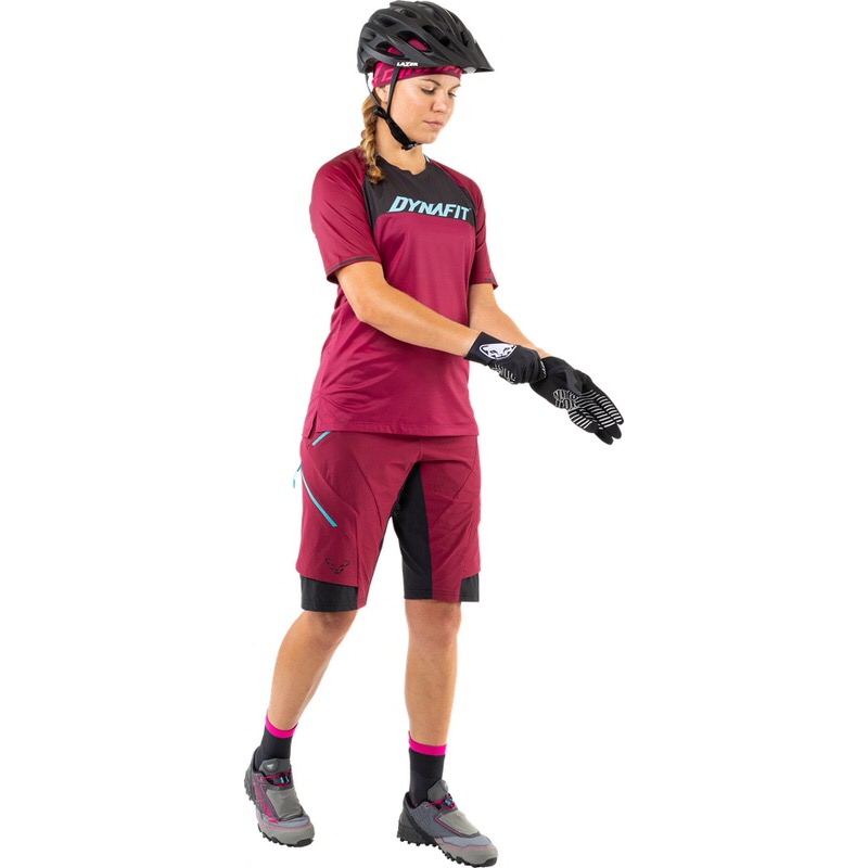 Frau in Beet Red Dynafit Ride Tee, Shorts, Helm und Handschuhen, beim Anziehen der Handschuhe.
